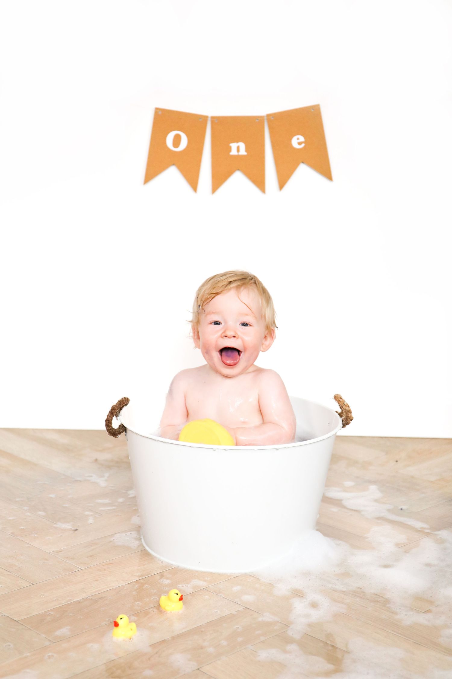 Happy baby sitting in white basin with rubber ducks under orange 'one' banner.
