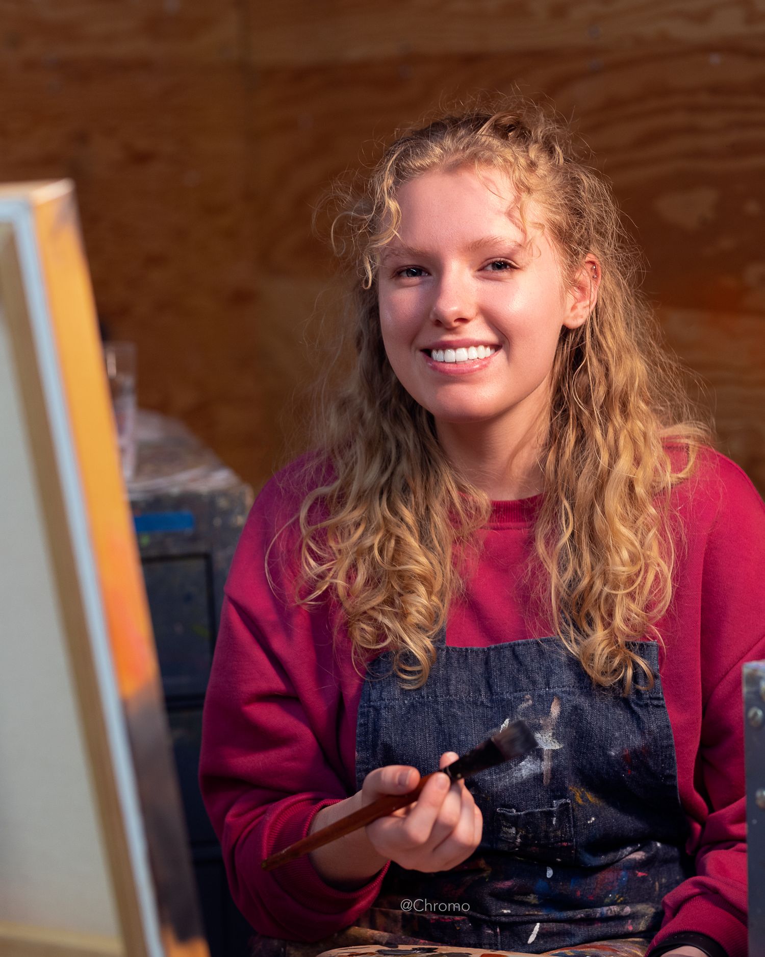 Portrait of an artist wearing overalls and holding painting supplies in a studio setting.