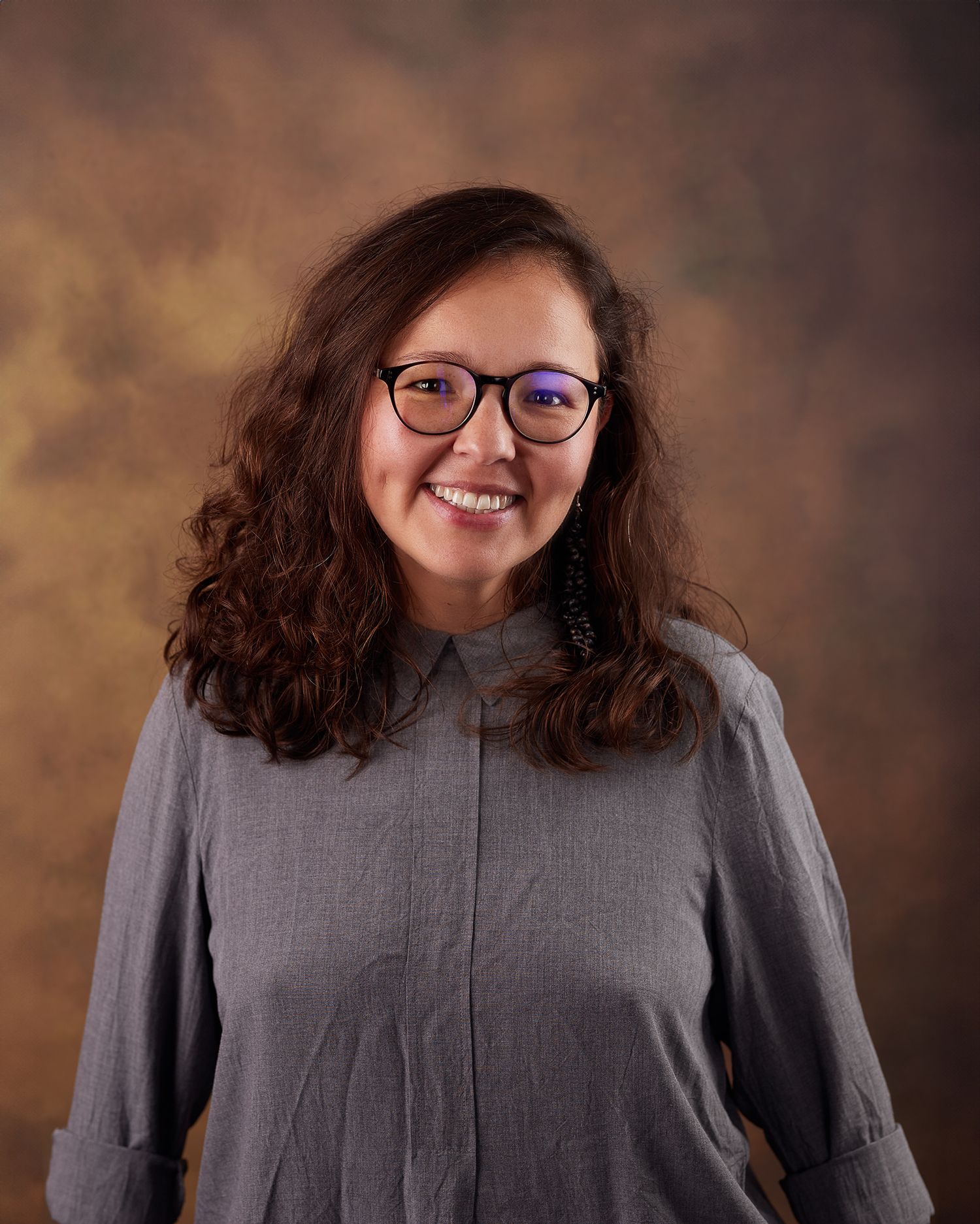 Professional headshot with purple glasses and grey button-up against warm backdrop.