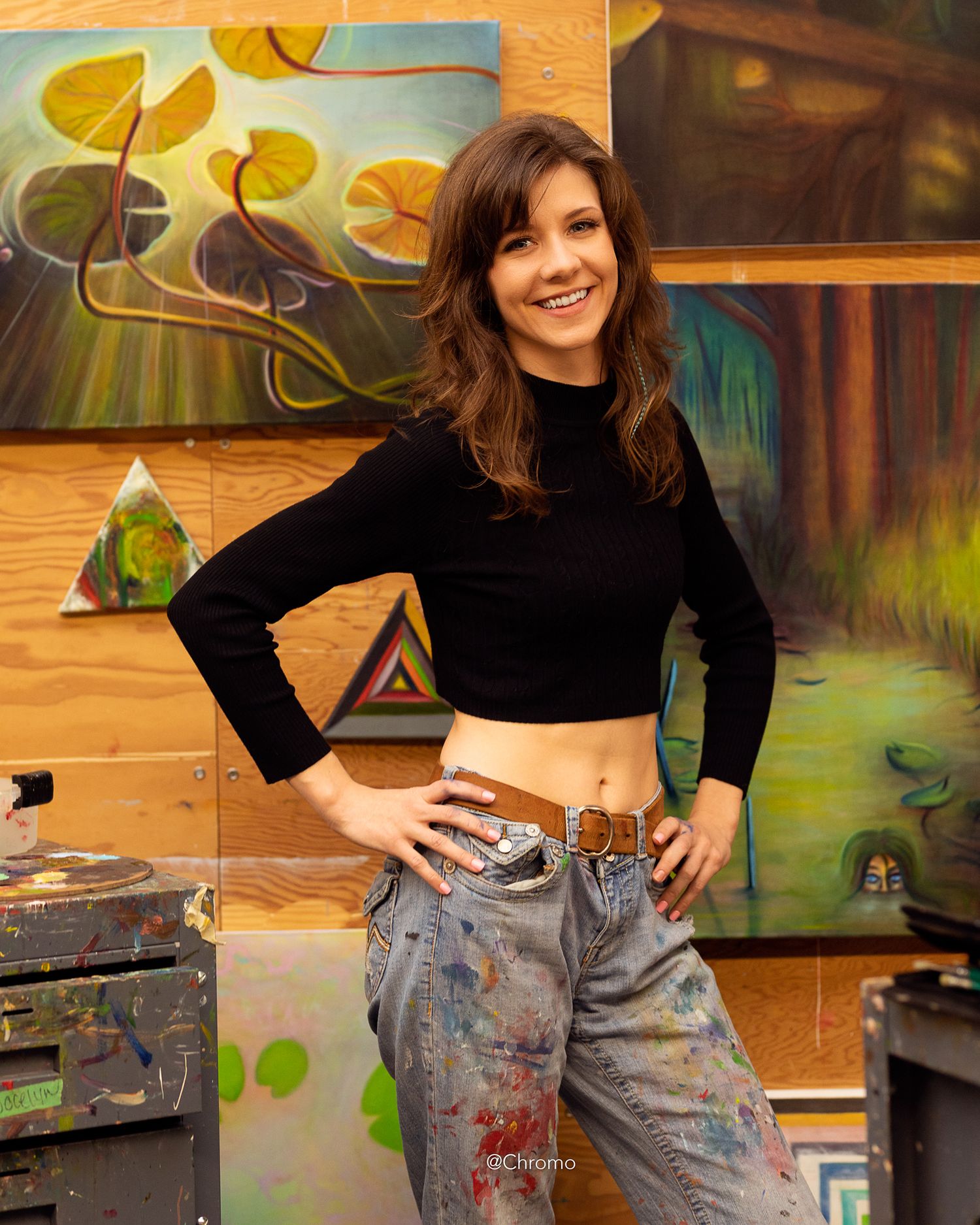 Artist in paint-splattered jeans posing in front of colorful artwork and paintings.