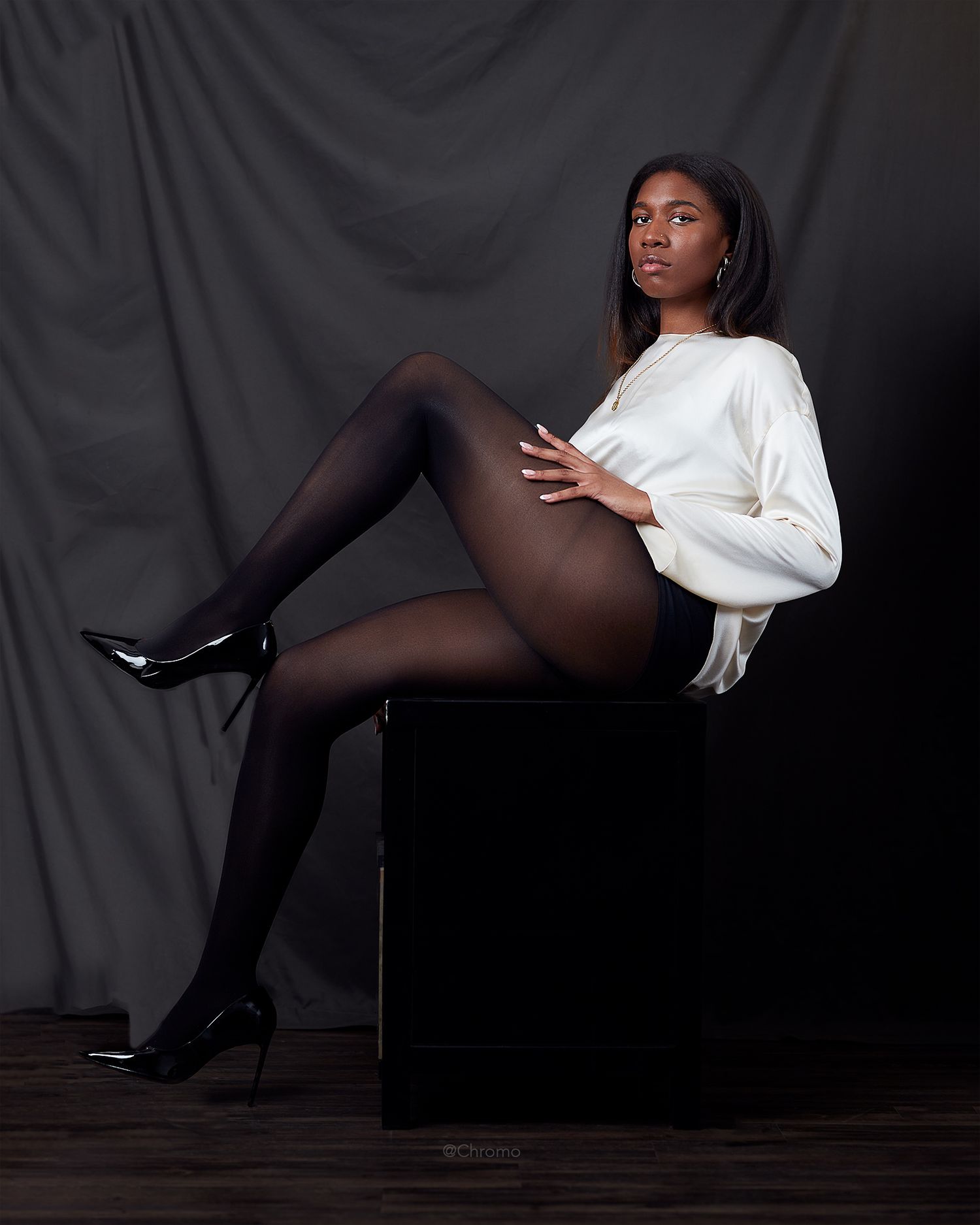 Fashion model in white sweater and black tights poses against dark background in artistic studio portrait.