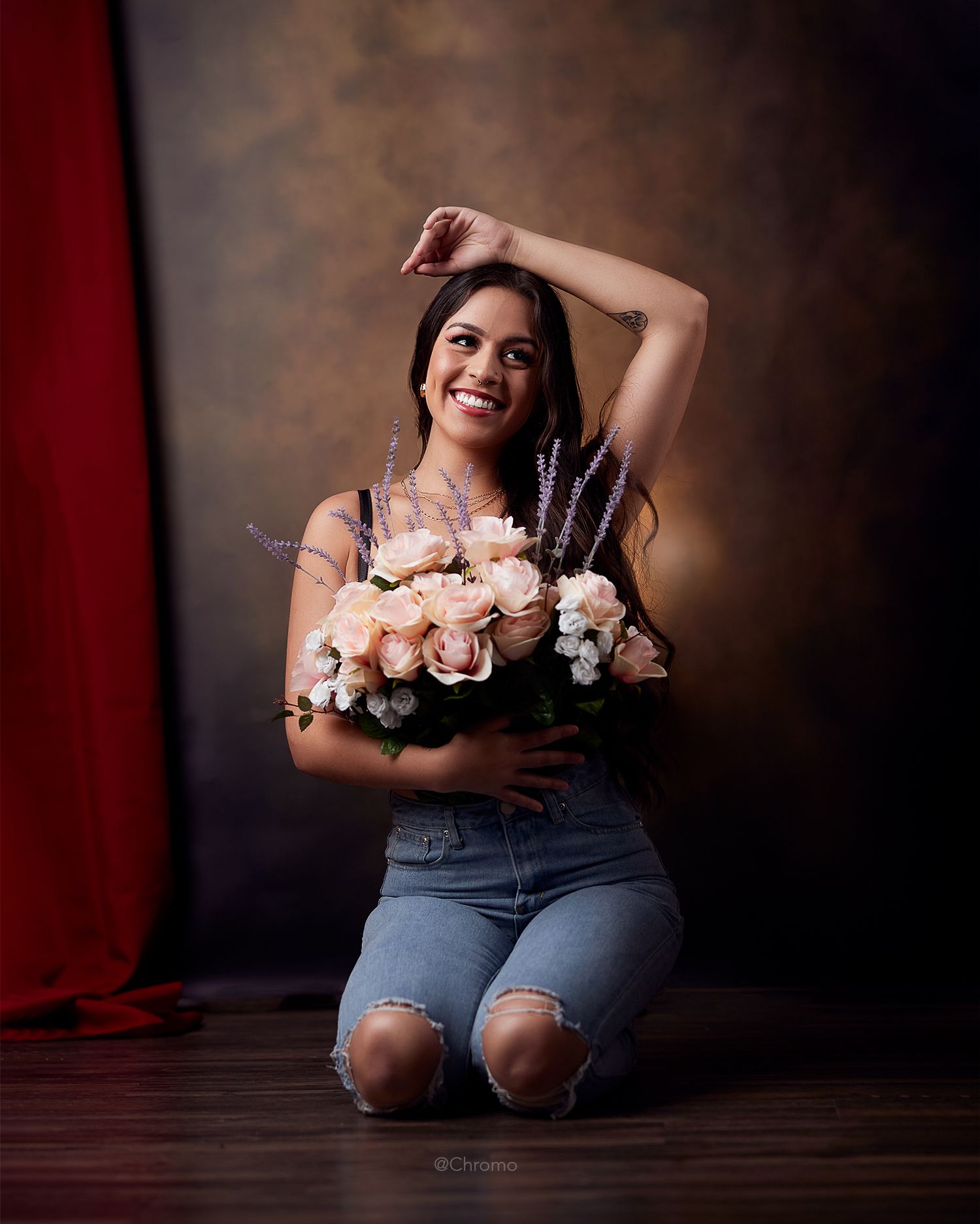 Portrait with bouquet of pink roses while kneeling on wooden floor against dark backdrop.