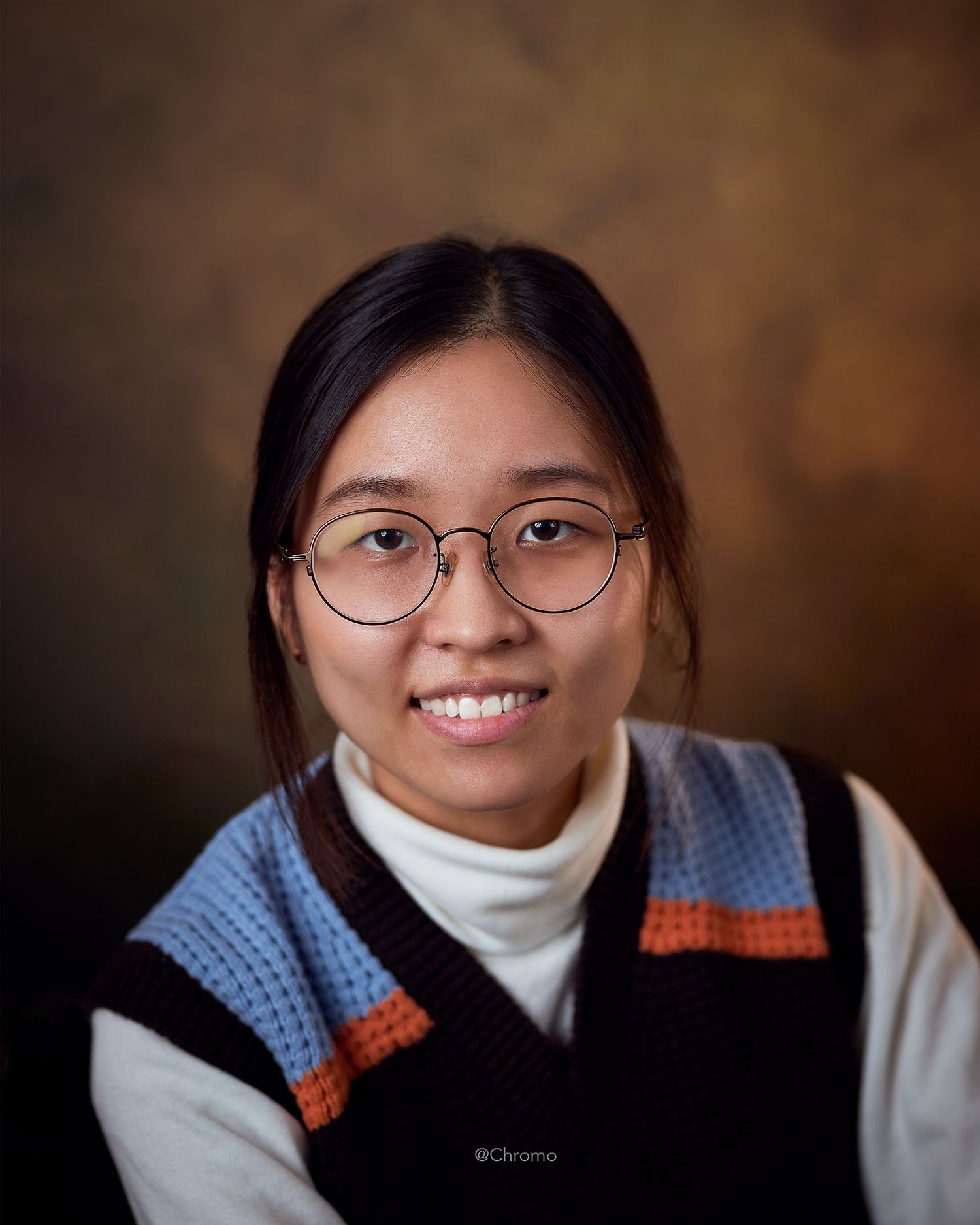 Portrait of person in glasses wearing knit vest and white turtleneck against brown background.