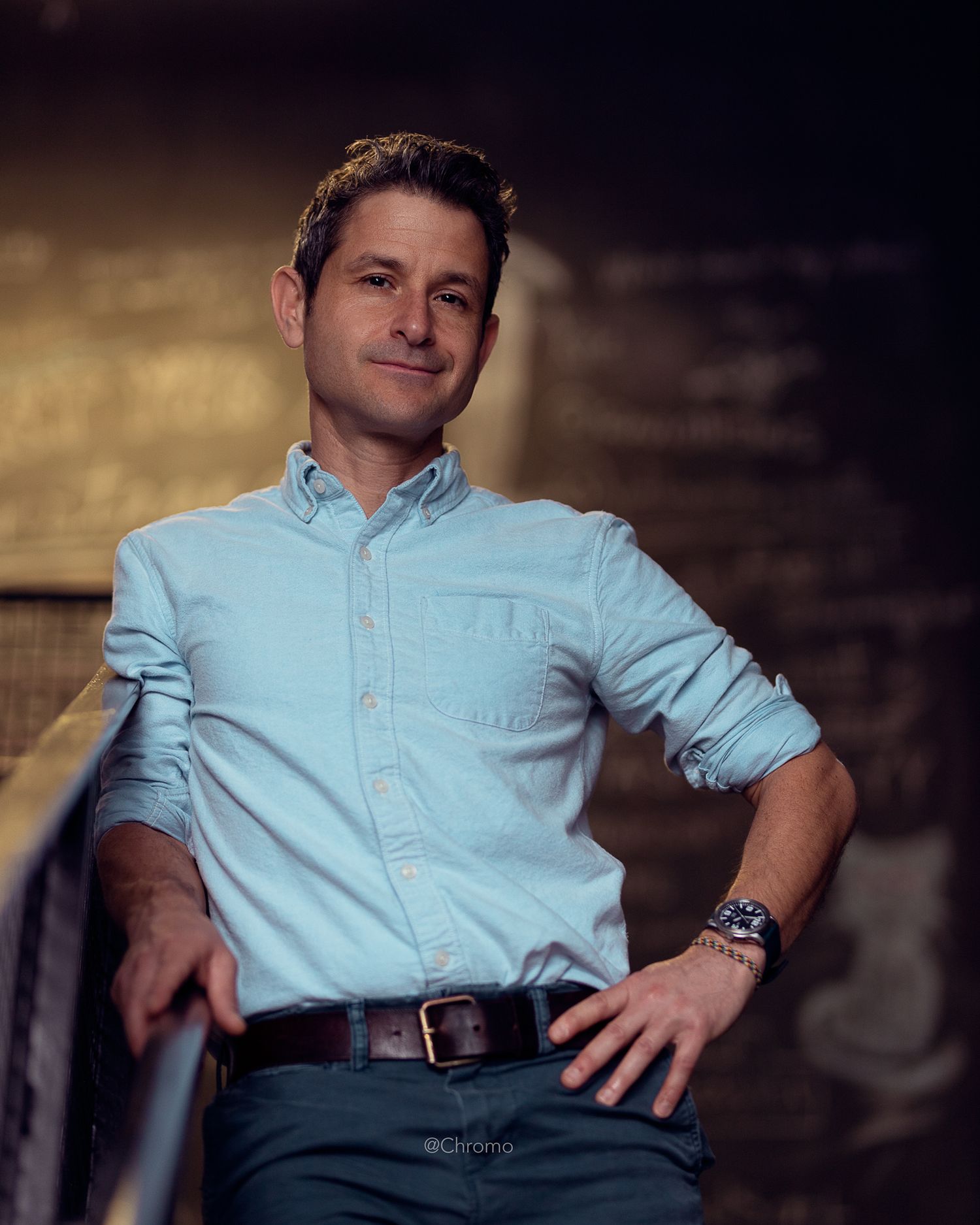 Portrait of person in light blue button-down shirt and dark pants against textured wall.