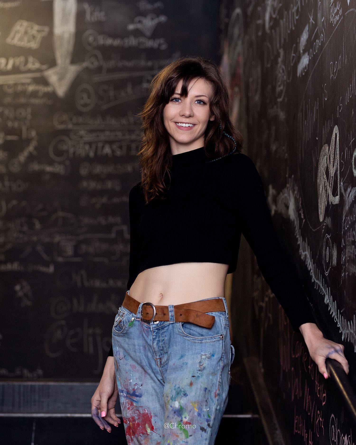 Portrait of someone in a crop top and paint-covered jeans against a wall with writing.