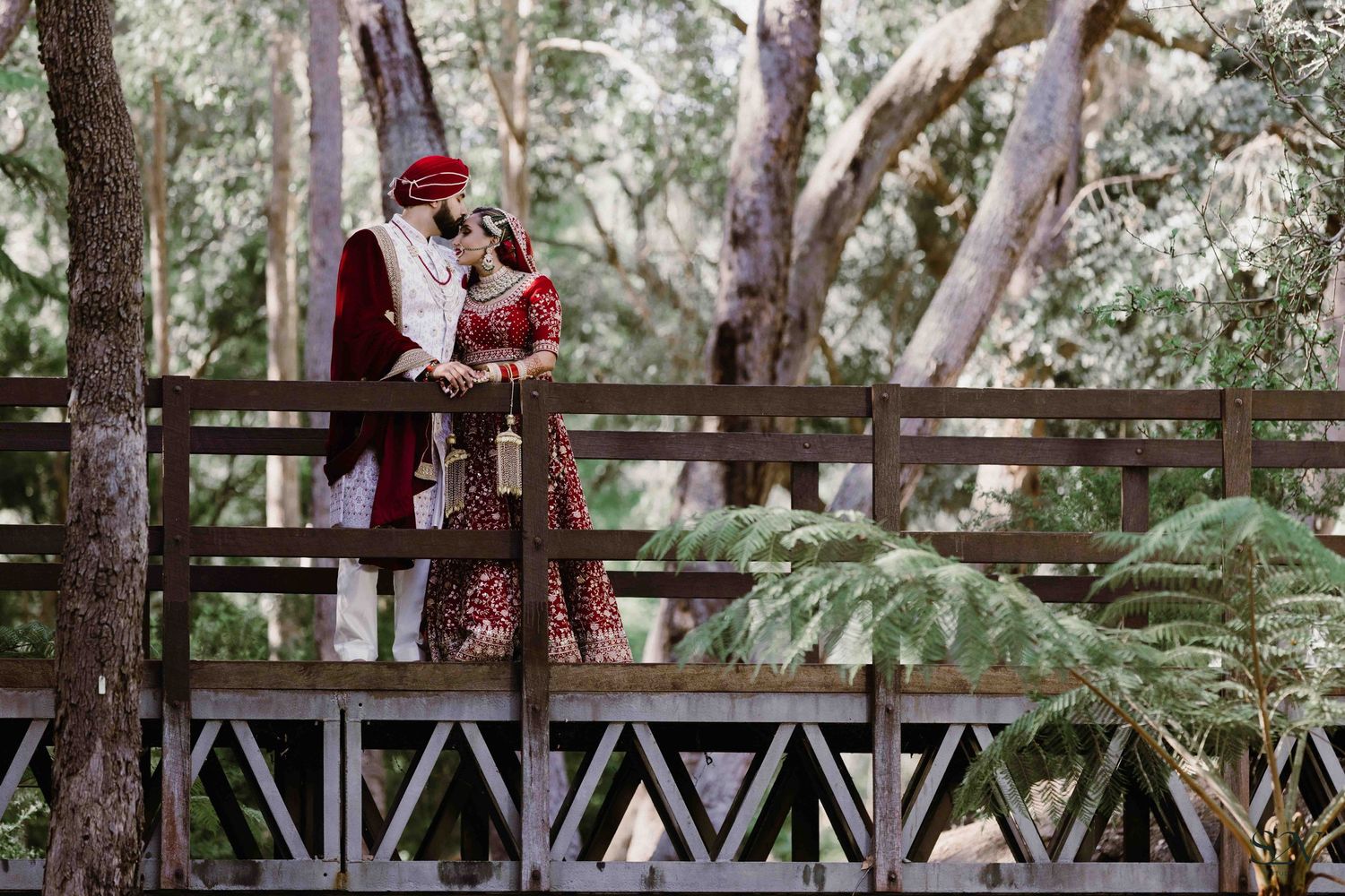 Couple in formal attire shares intimate moment on wooden bridge surrounded by lush forest.