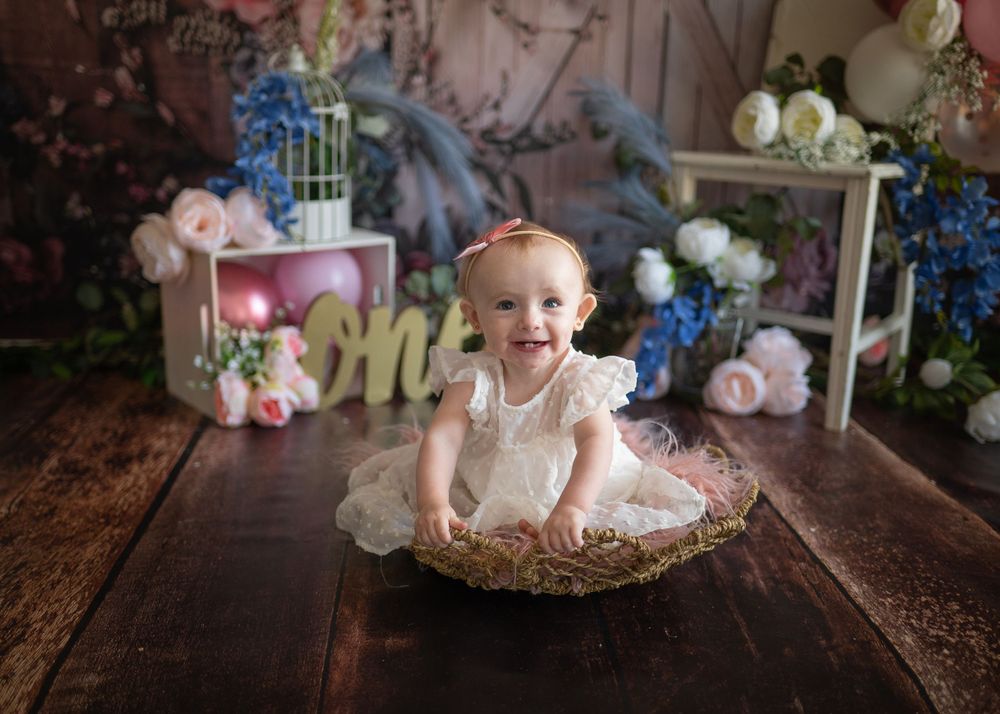 Baby in white dress sits in decorative nest with blue and pink party decorations.