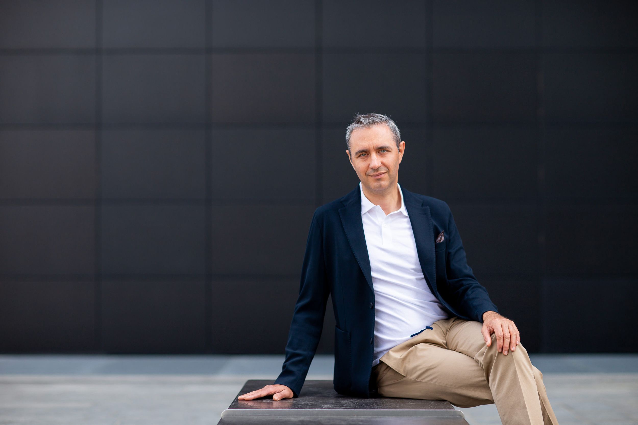 Professional portrait of person in navy blazer sitting casually against dark wall.