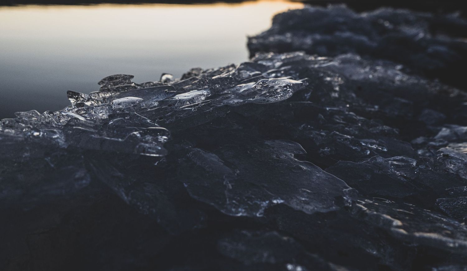 Dark moody close-up of rough icy rocks at waters edge with reflective surface in background.
