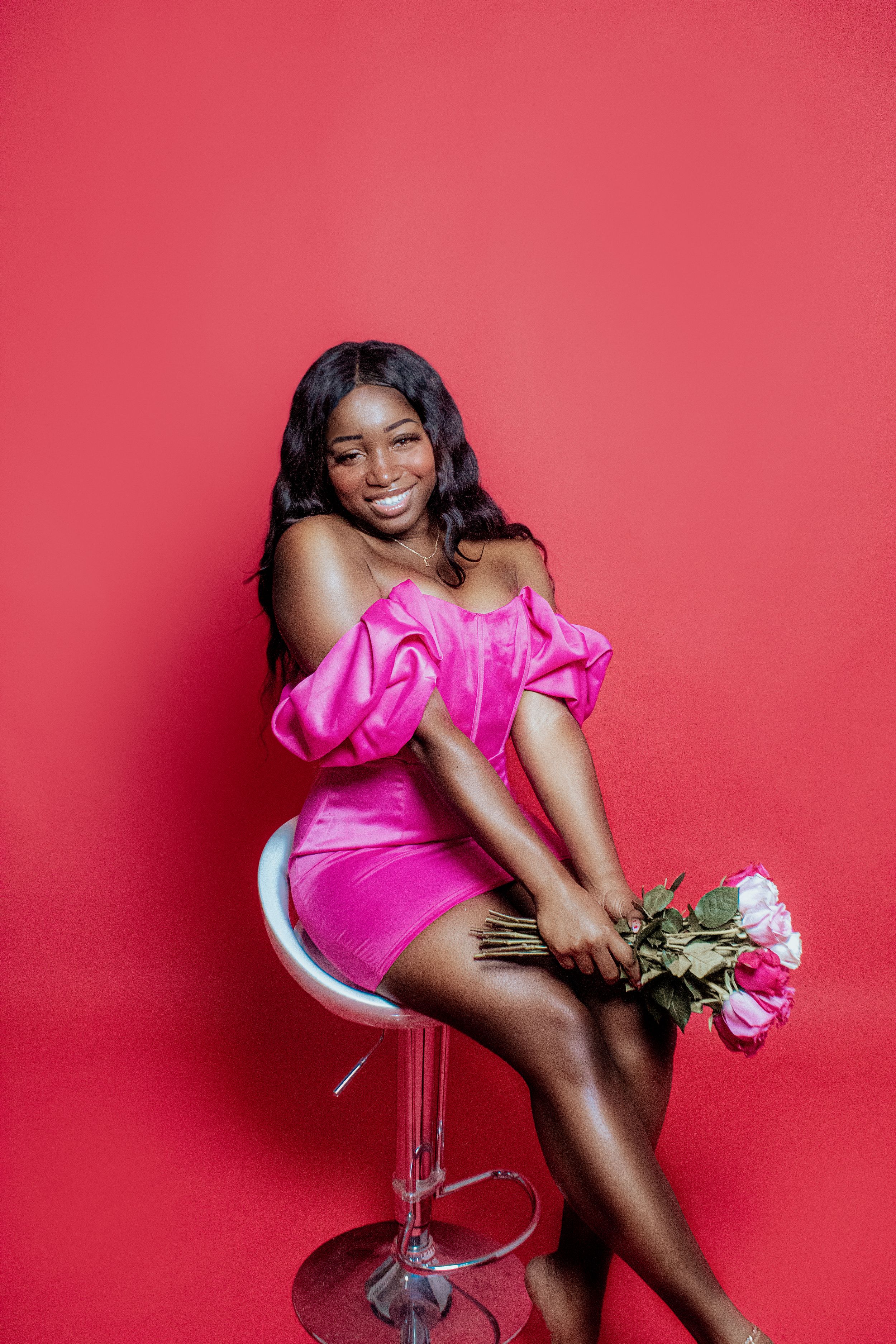 Elegant model in hot pink off-shoulder dress poses on silver barstool against red backdrop.
