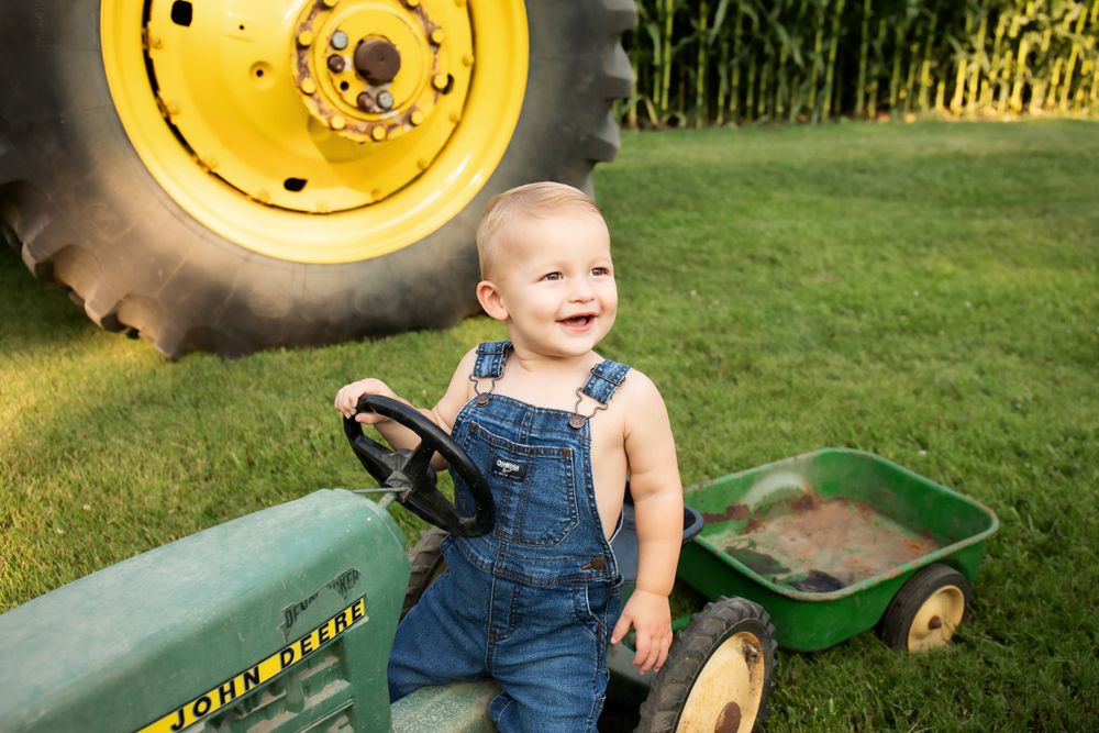Joyful toddler in overalls playing on green toy tractor beside large tractor wheel.