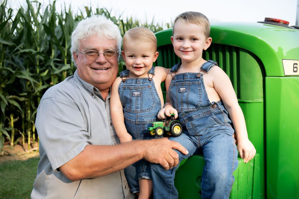 Three generations pose together by antique green tractor in rural setting.
