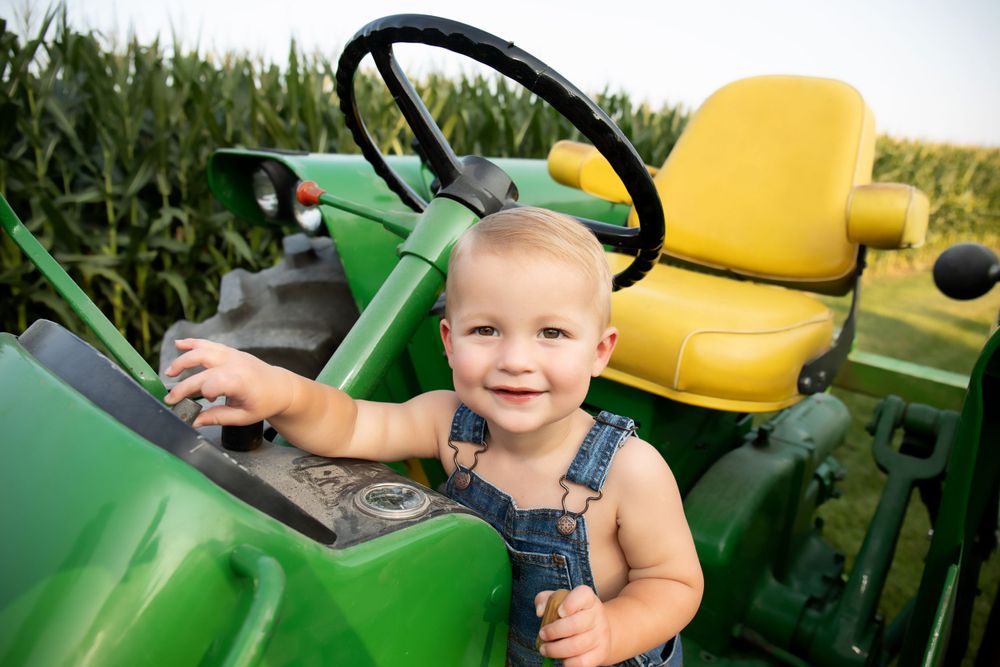 Happy toddler in denim overalls sits in a John Deere tractor with yellow seat overlooking cornfield.