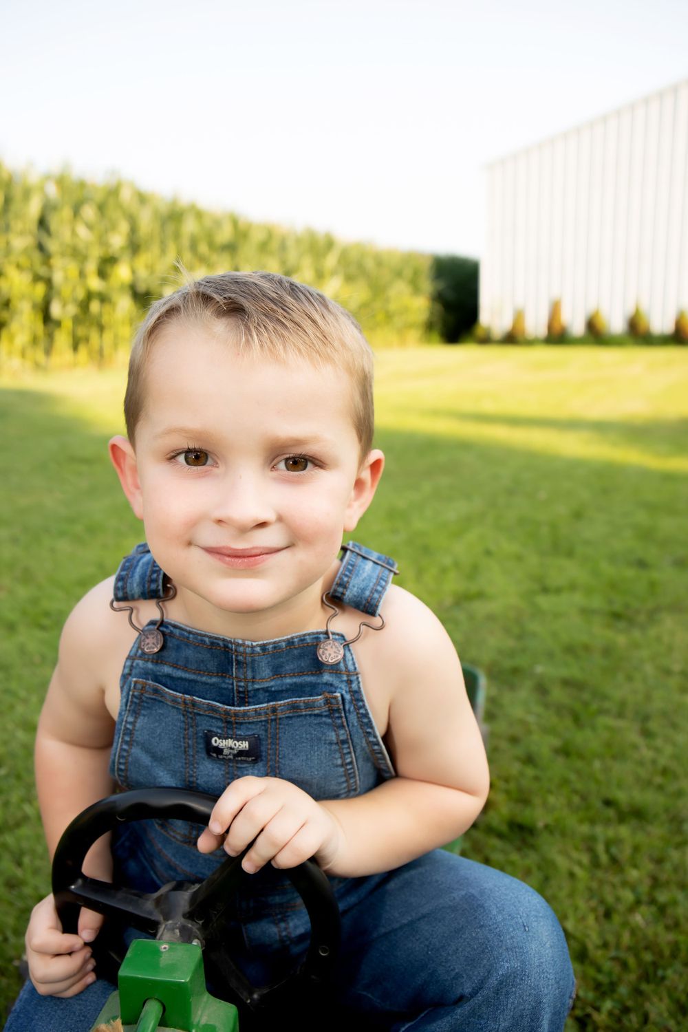 A young child in denim overalls sits on a toy tractor in a sunny field.