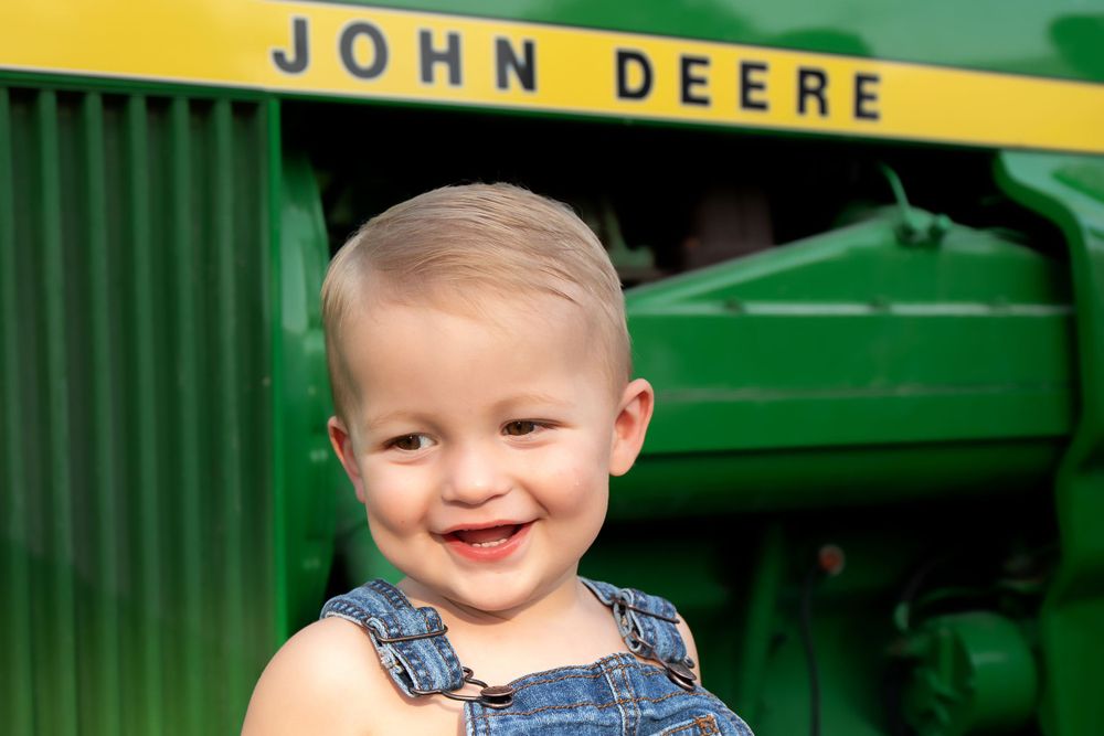 Smiling child in overalls poses in front of green John Deere tractor.