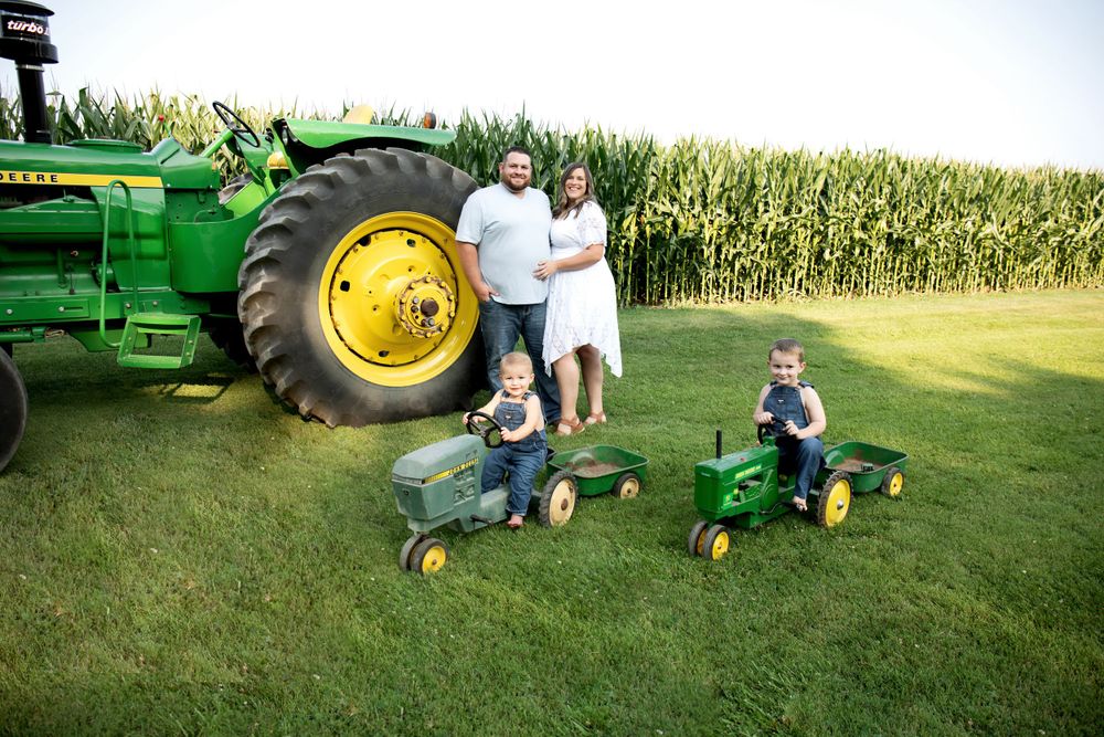 Family poses with John Deere tractor and toy tractors on grass near cornfield at sunset.