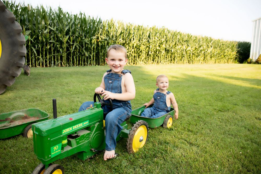 Two children playing on toy John Deere tractors in grassy field near cornfield.