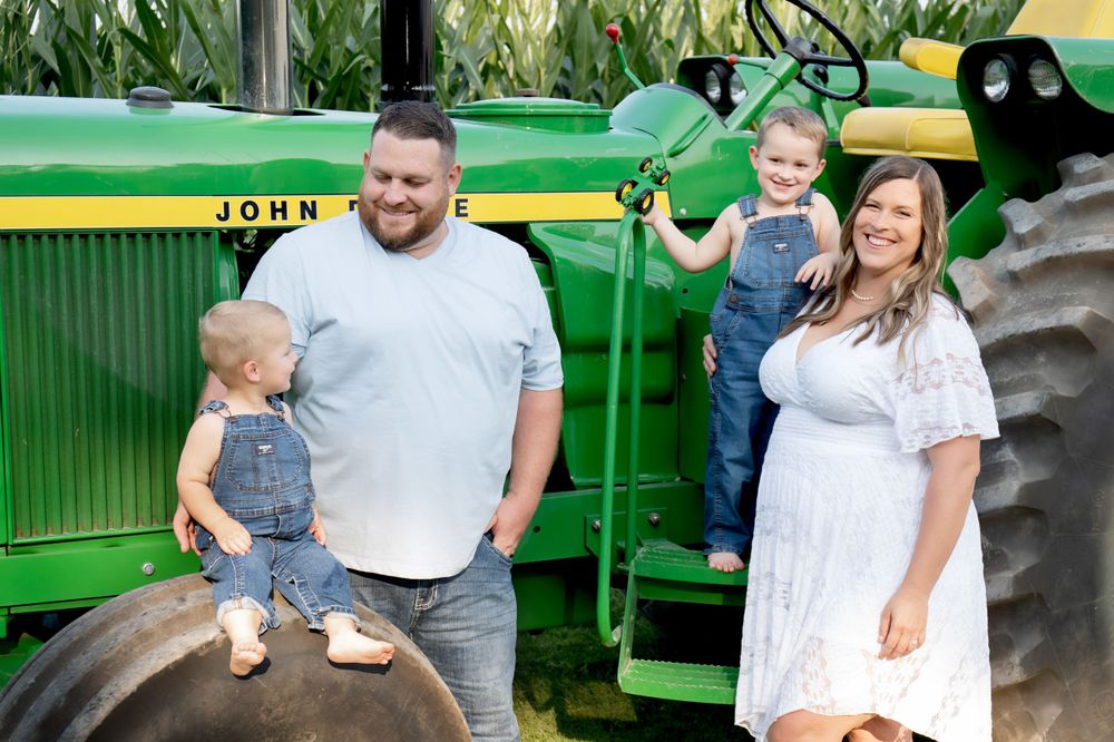 Family in casual attire poses together beside large green John Deere tractor.