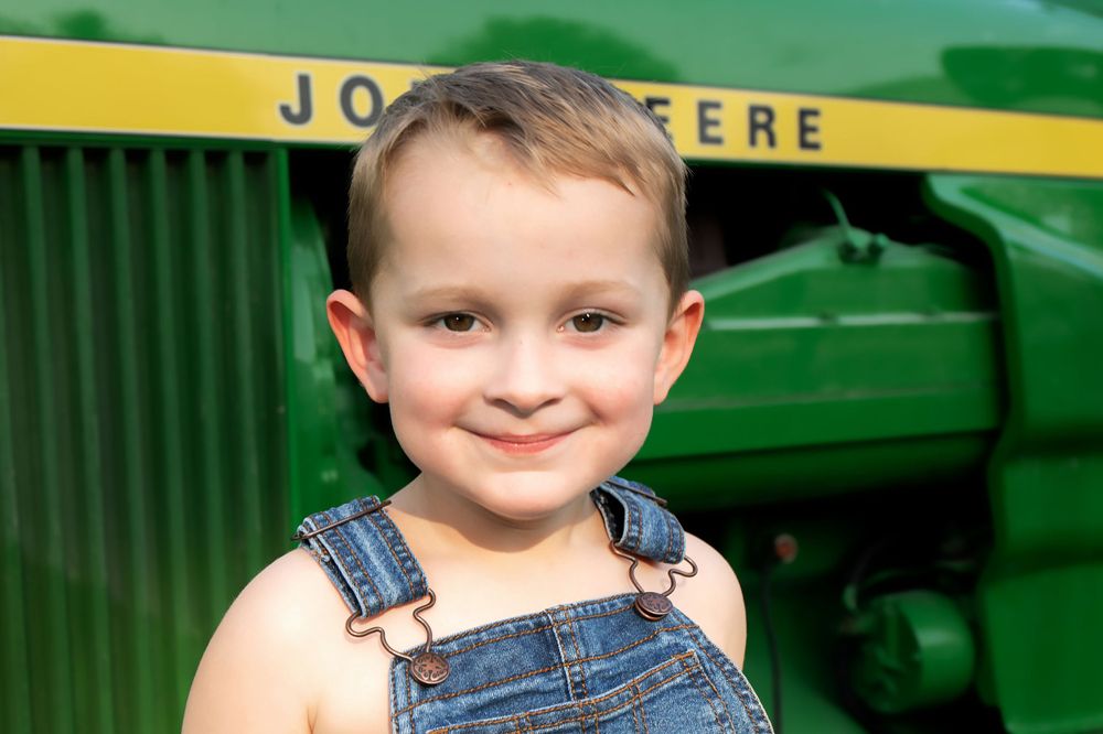 Child in denim overalls poses by vintage John Deere tractor.