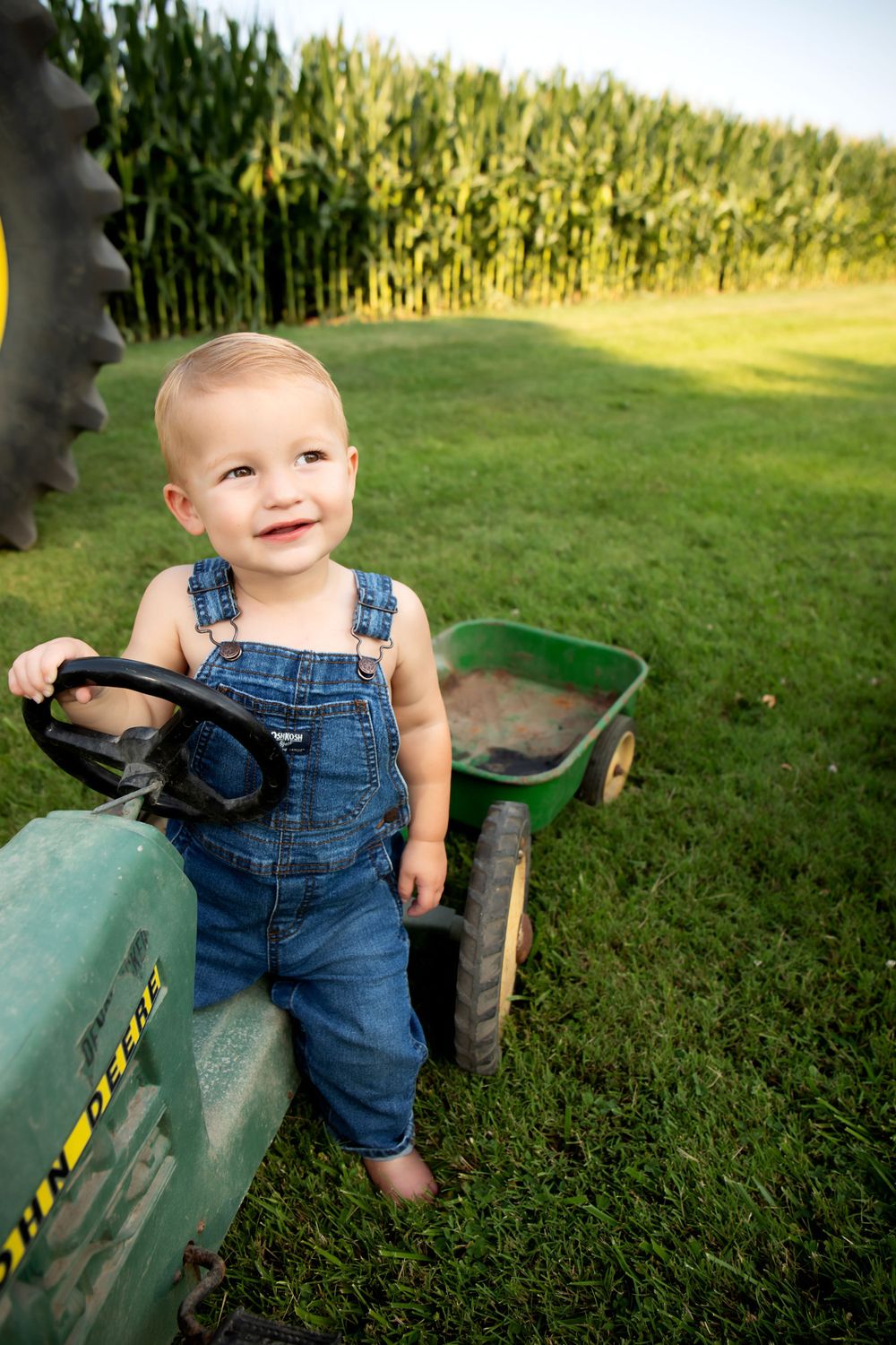 Small child in denim overalls stands by vintage John Deere tractor against cornfield backdrop.