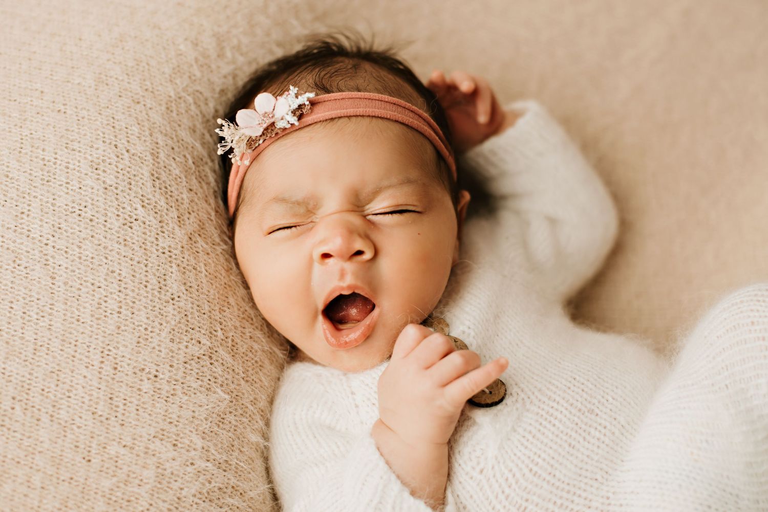 Sleeping newborn wearing a delicate floral headband and white sweater yawns while resting.
