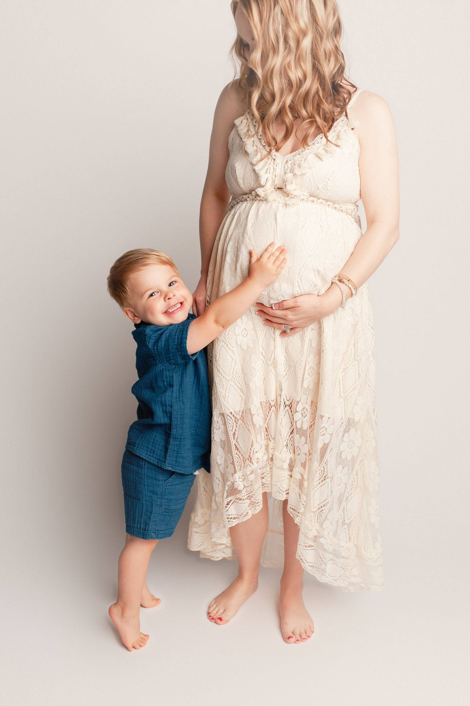 Smiling toddler boy in blue outfit hugging pregnant mother in white lace dress.
