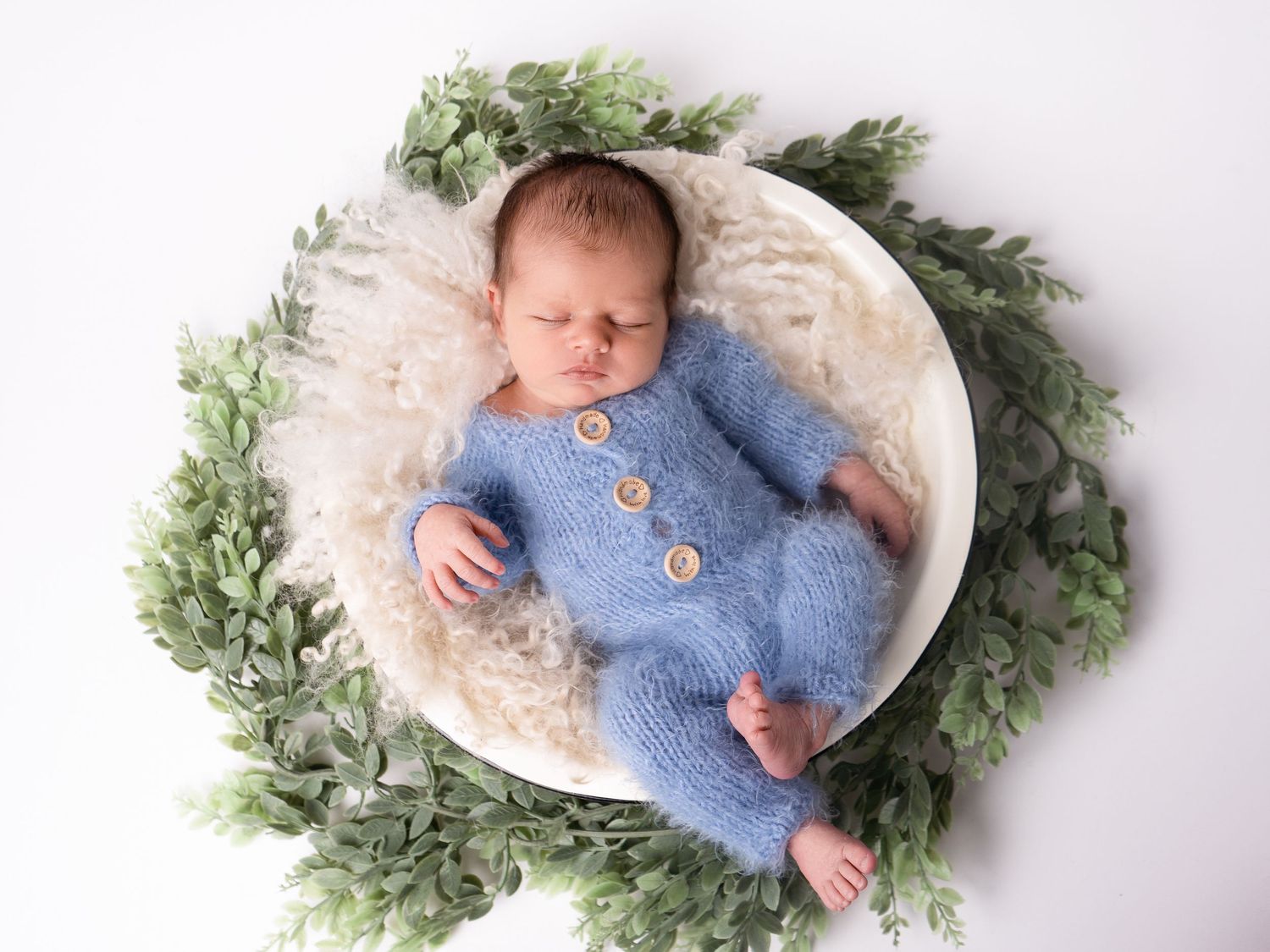 Sleeping newborn baby boy in a white bowl with greenery, wearing a blue fluffy outfit at newborn photoshoot in Doncaster