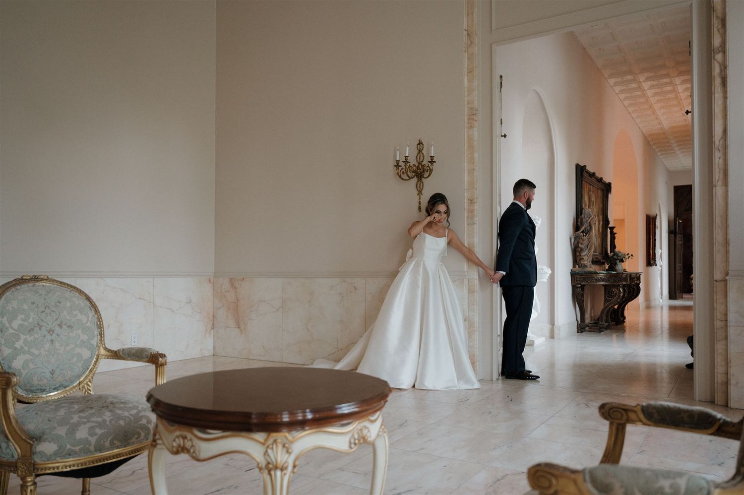 A couple stands apart in an elegant white hallway with vintage furniture and a chandelier during a formal occasion.