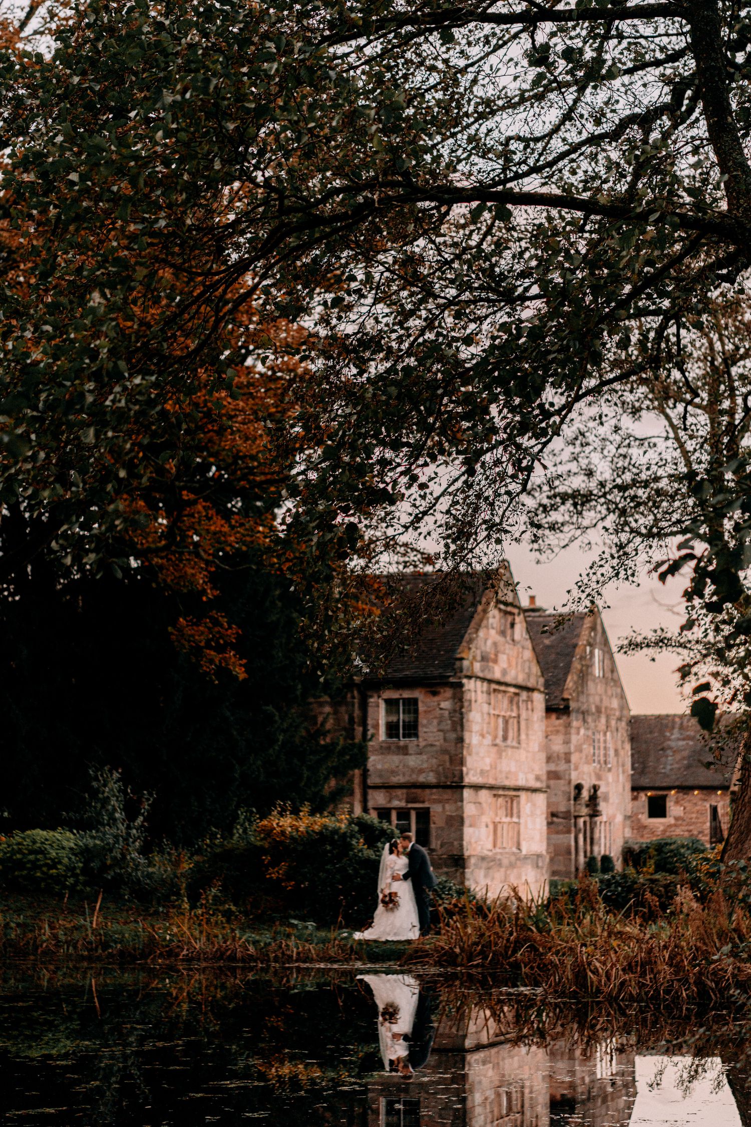 A moody autumn scene shows an old stone manor house reflecting in a pond as leaves turn orange at dusk.