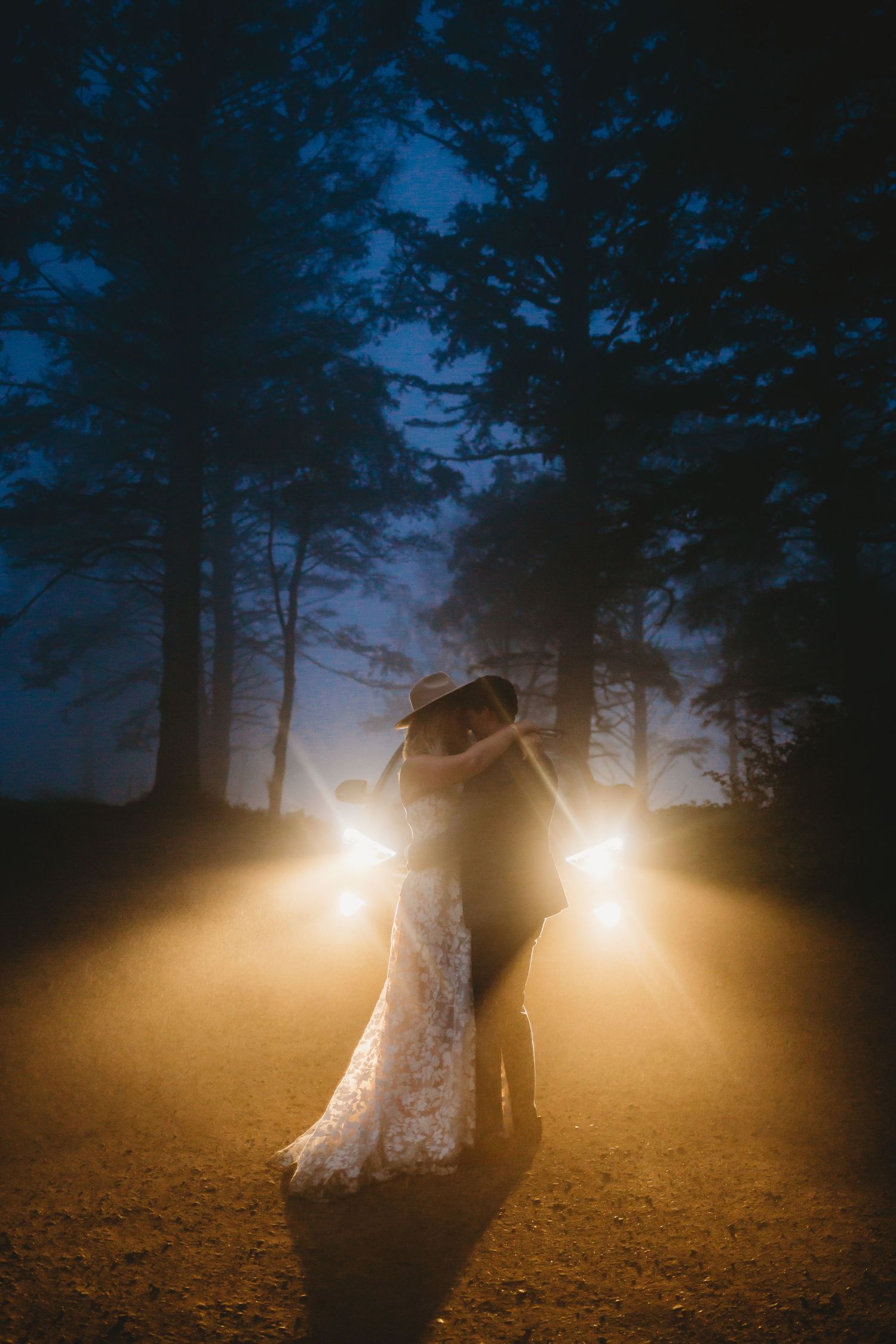 A bride and groom are sharing their first dance during blue hour in the light of their car's headlights.
