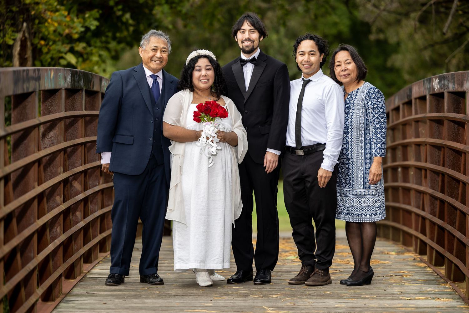 A family group photo on a wooden bridge with the bride in white holding red roses.