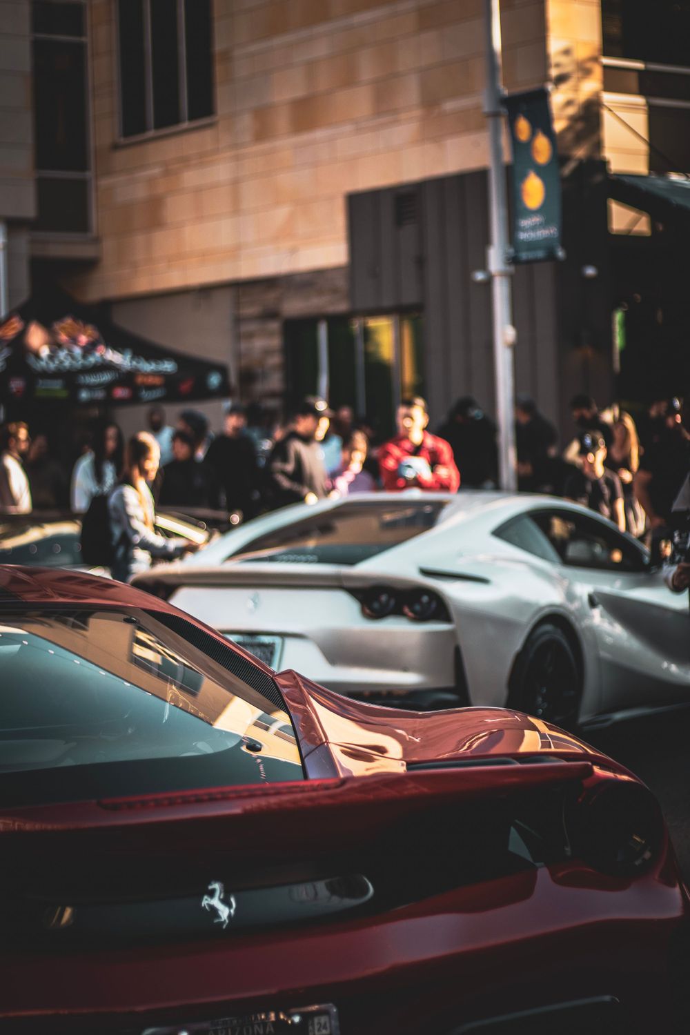 Luxury sports cars including a red Ferrari parked on a city street with people gathered nearby.
