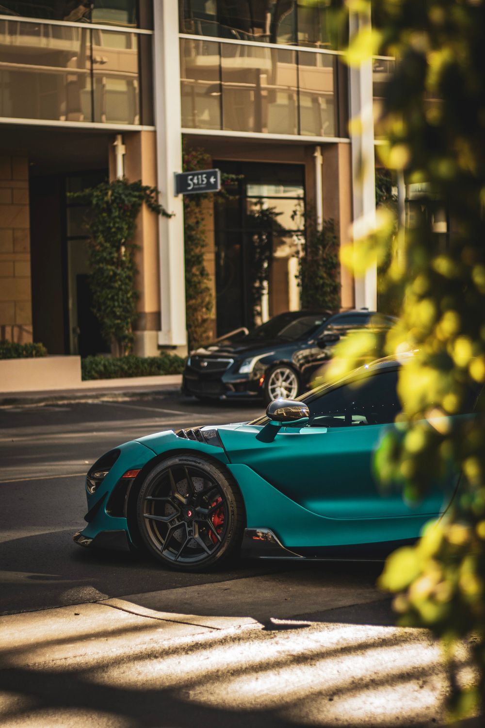 Teal McLaren supercar parked on a street with modern architecture and foliage in the background.