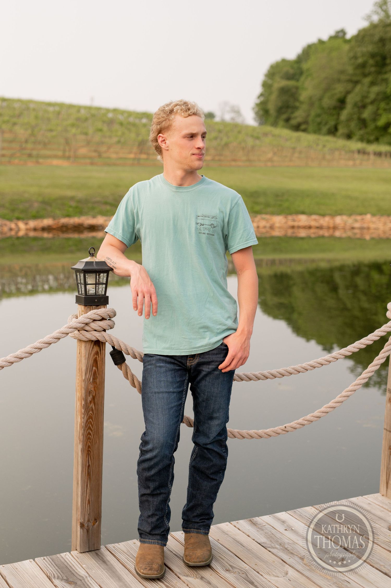 Person in mint t-shirt stands on wooden dock with lantern post by lake.
