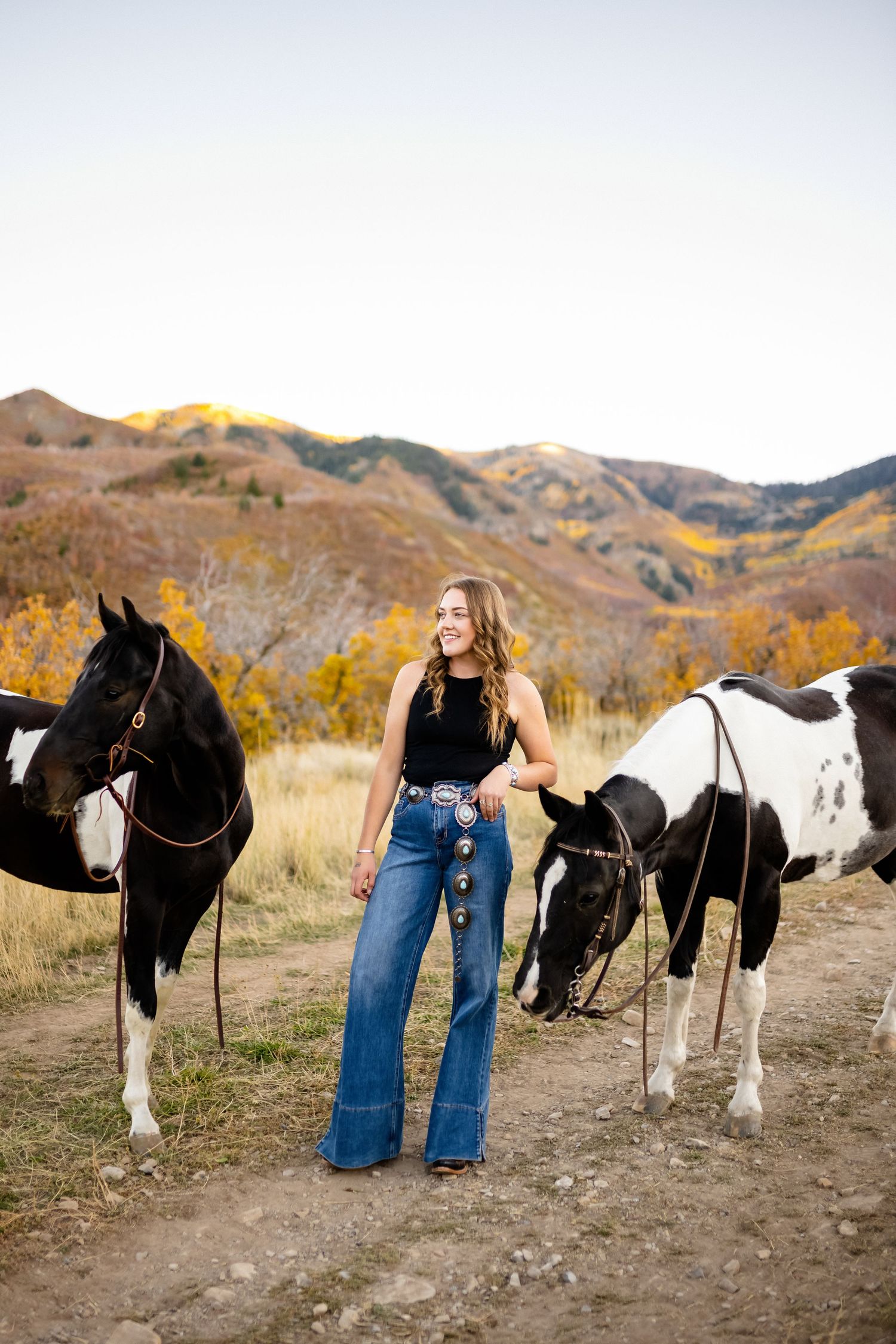 A woman stands between two paint horses against a backdrop of fall-colored mountains.