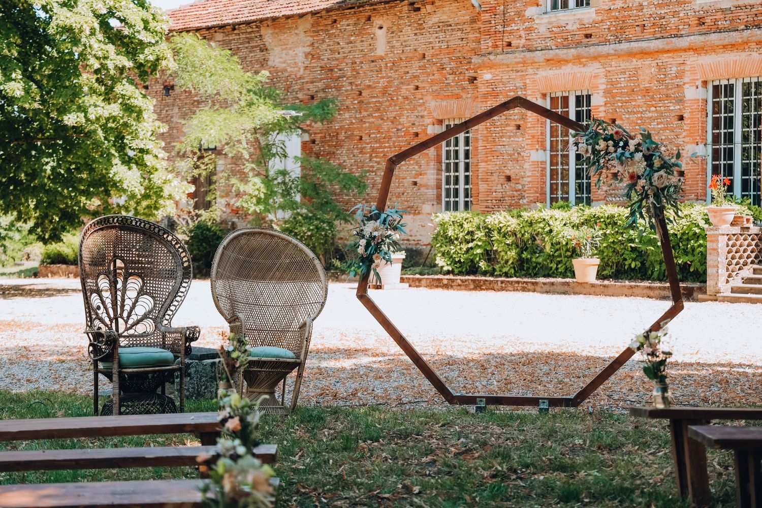Rustic outdoor wedding arch with wicker chairs set up against brick building backdrop.