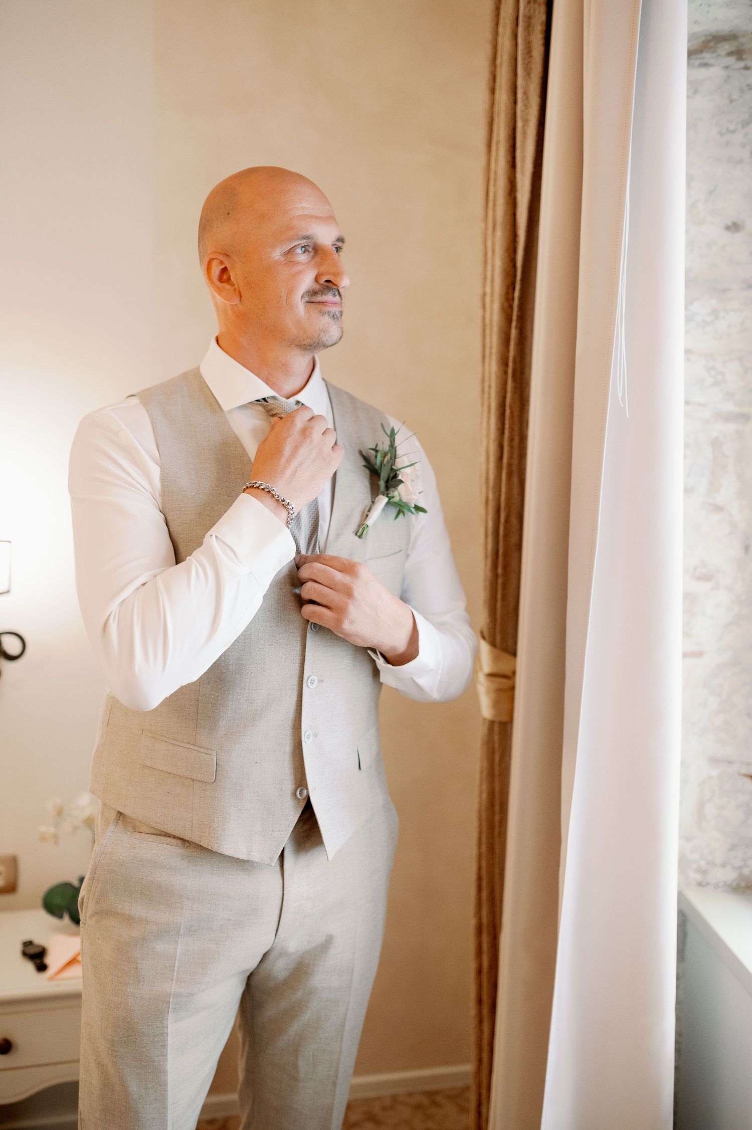 A groom in a beige suit vest adjusts his tie while standing by a window.