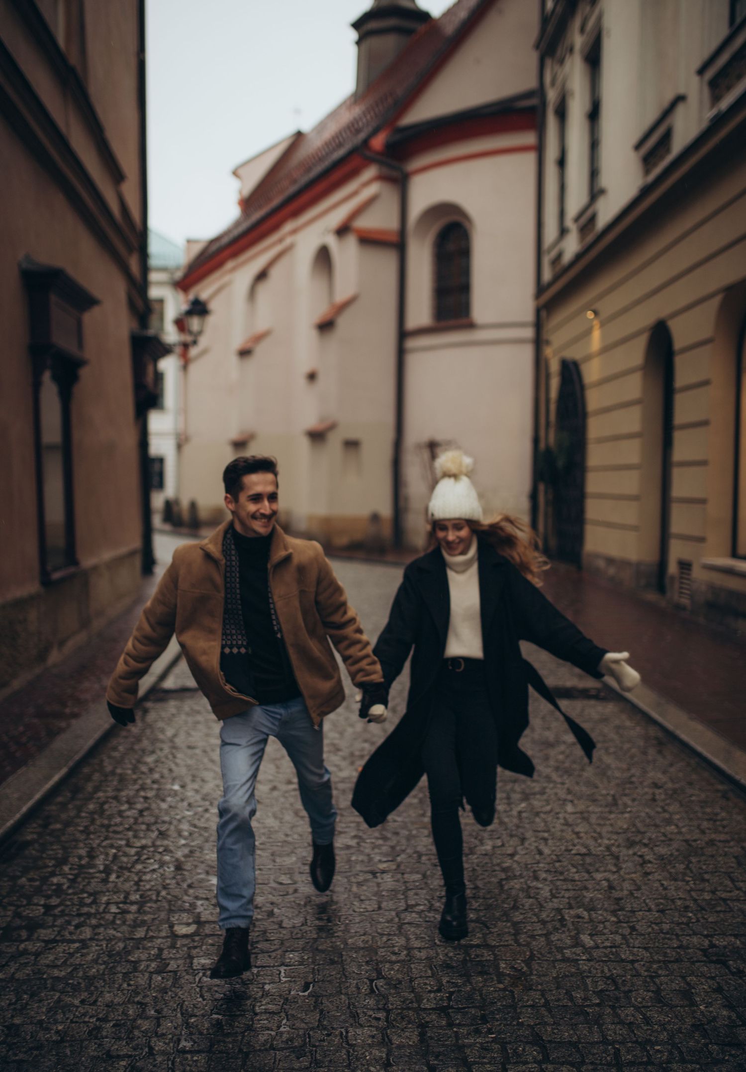 Playful couple running down cobblestone street with historic architecture in background.