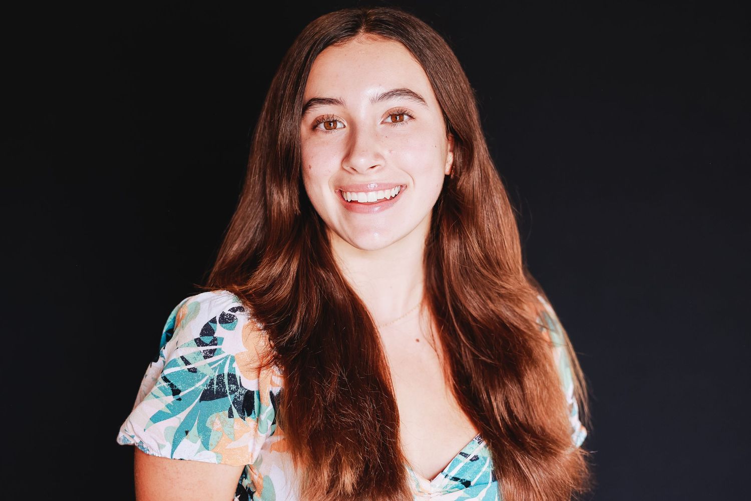 Cheerful portrait in floral dress with long brown hair against black backdrop.
