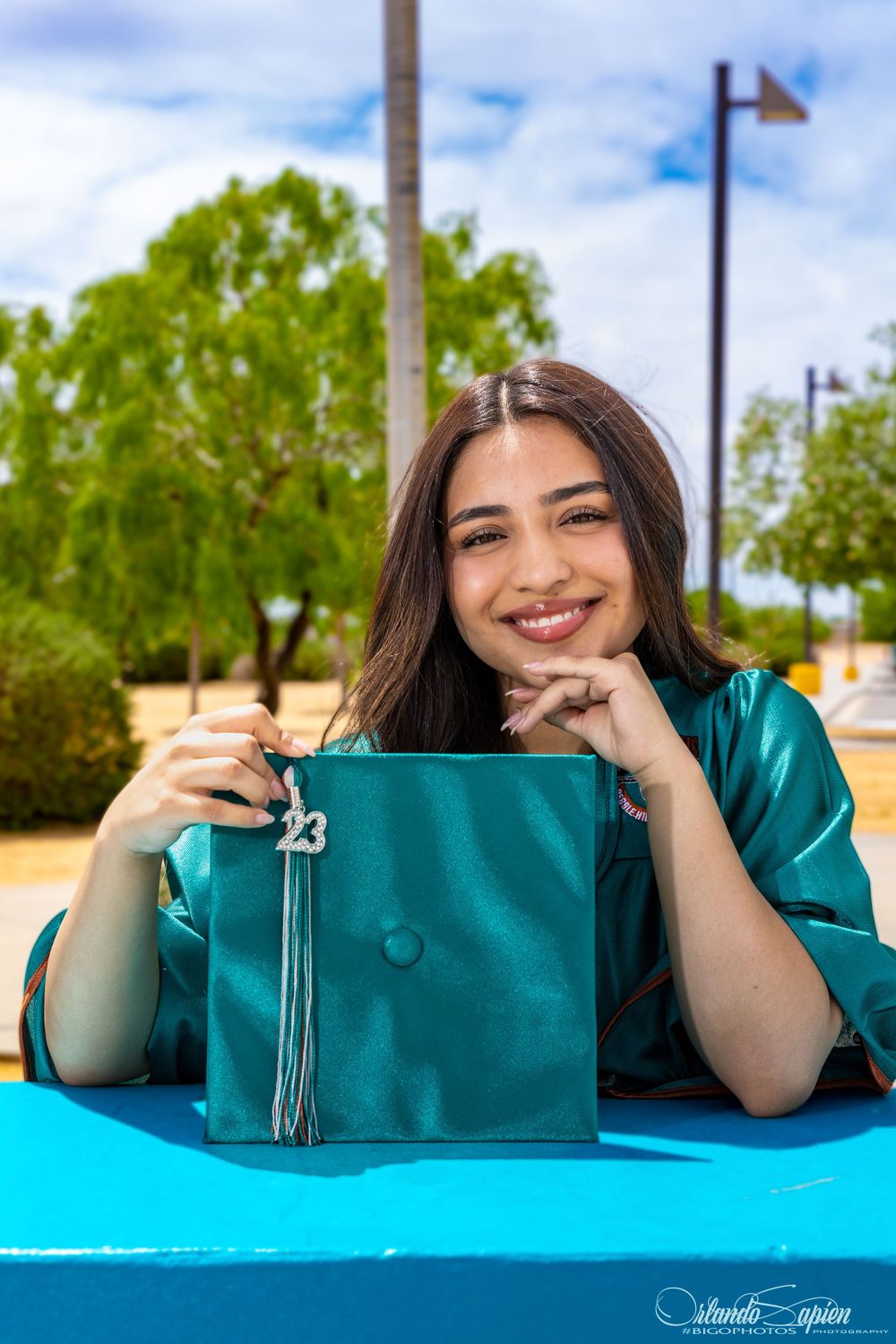 A teal handbag displayed on a bright blue surface outdoors with trees and sky in the background.