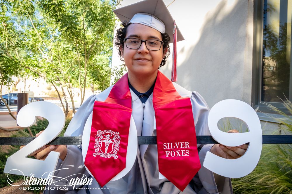 Graduate wearing white gown and red stole poses for graduation photos in sunny outdoor setting.