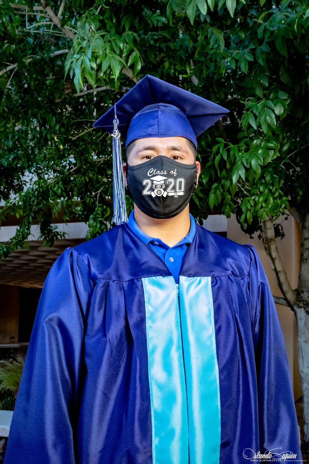 Graduate wearing blue cap and gown with face mask poses outdoors for graduation photos.