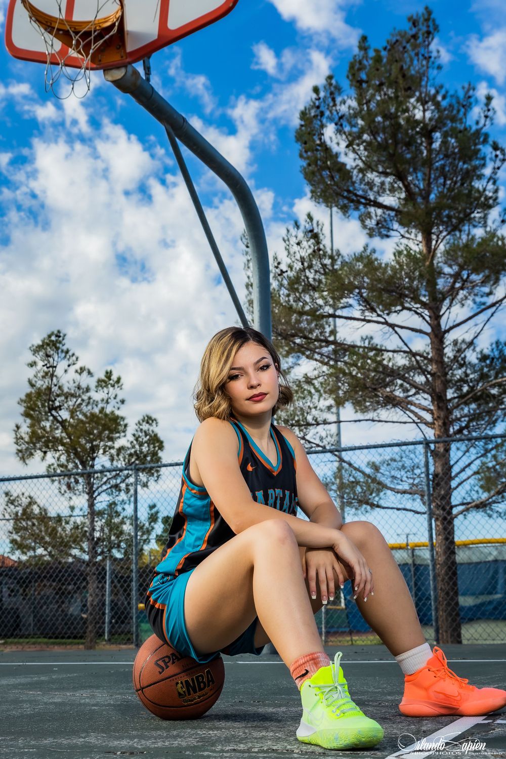 Basketball player in teal uniform sits on court with basketball near hoop.