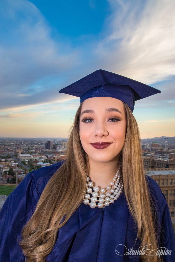Graduate in navy cap and gown poses against city skyline at sunset.