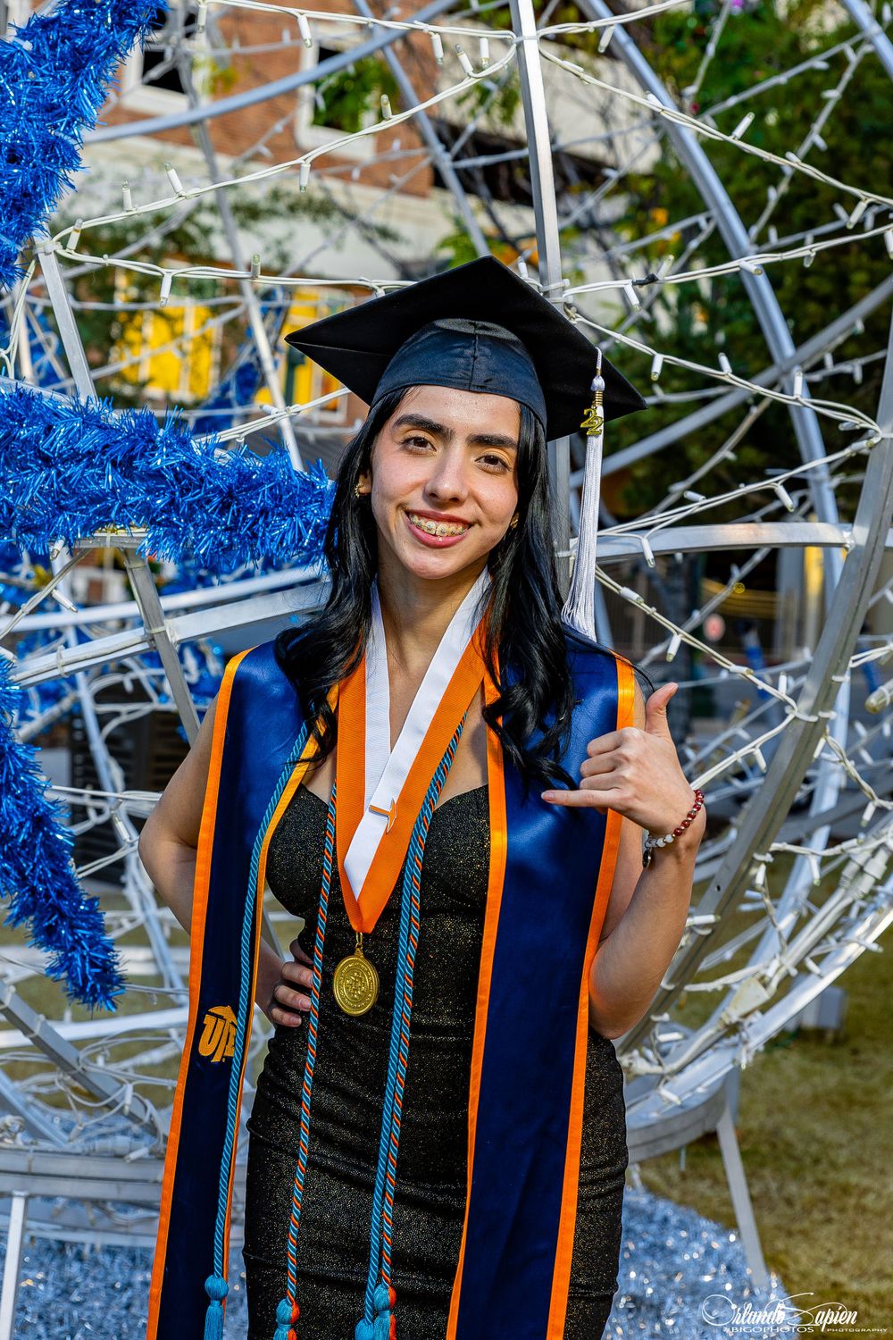 Graduate in cap and gown poses with decorative blue and silver backdrop.