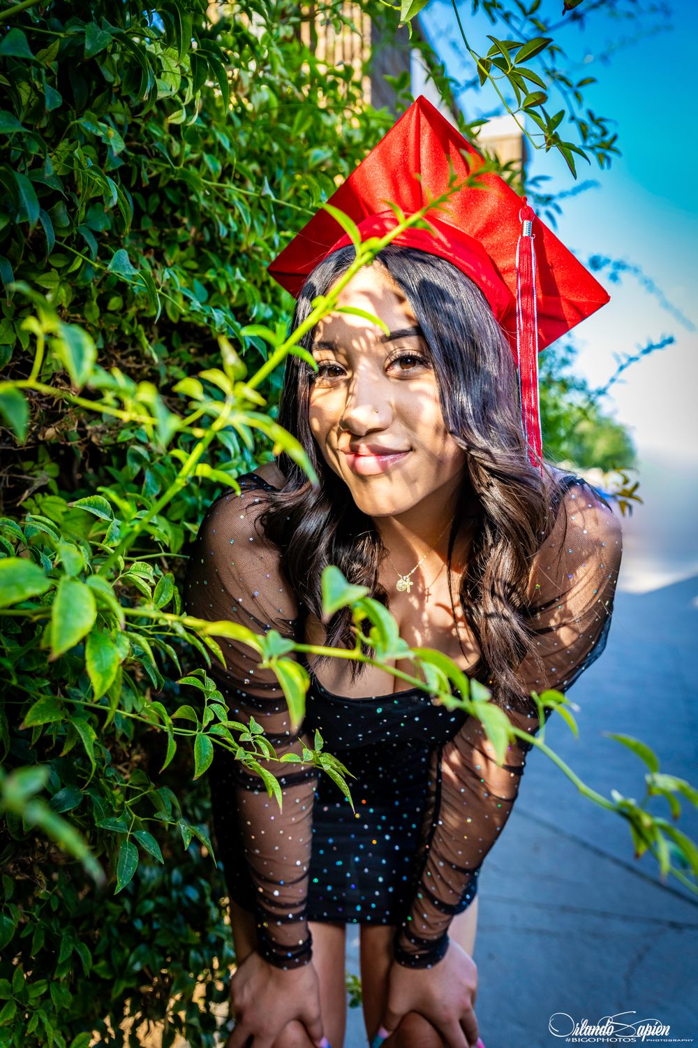 Graduate in red cap and black dress poses playfully among green foliage.