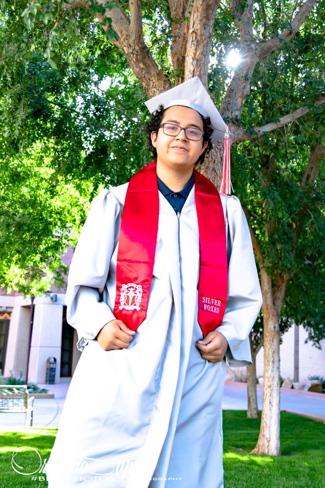 Graduate in white gown and red stole poses outdoors in front of trees during graduation photo shoot.