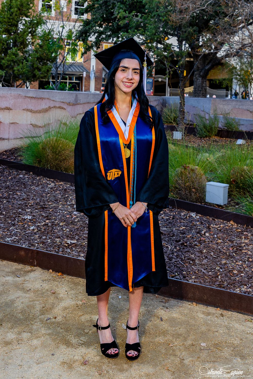 Graduate in blue and orange academic regalia poses outdoors.