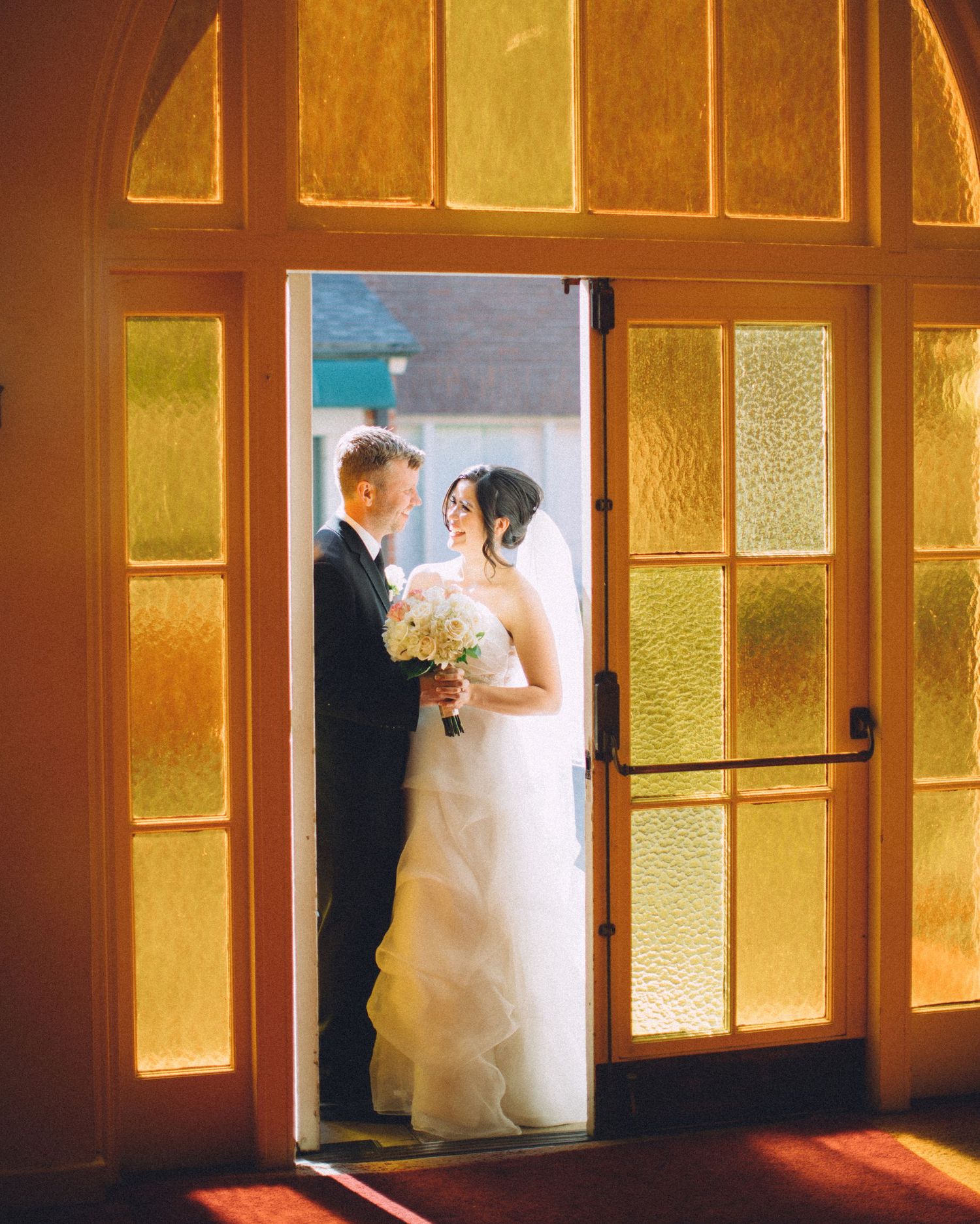 Cinematic wedding portrait of a bride and groom sharing a quiet moment framed by warmly lit amber glass doors.