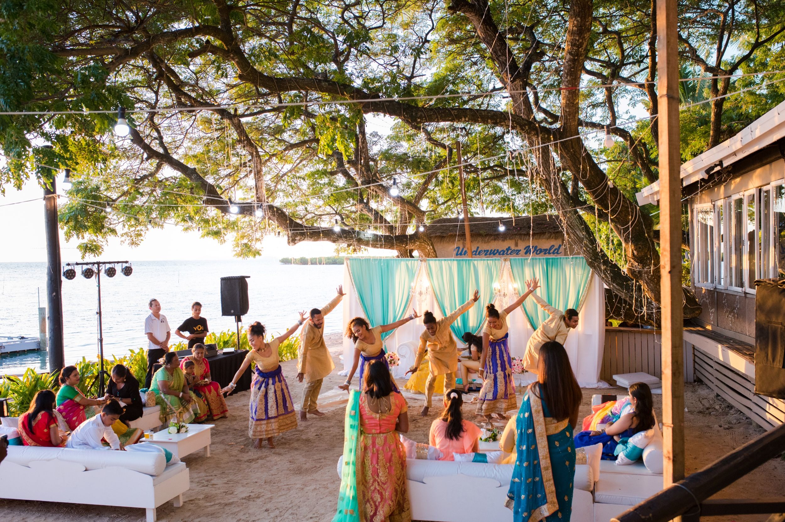 Joyful guests dance together at an outdoor celebration under trees with turquoise drapes.